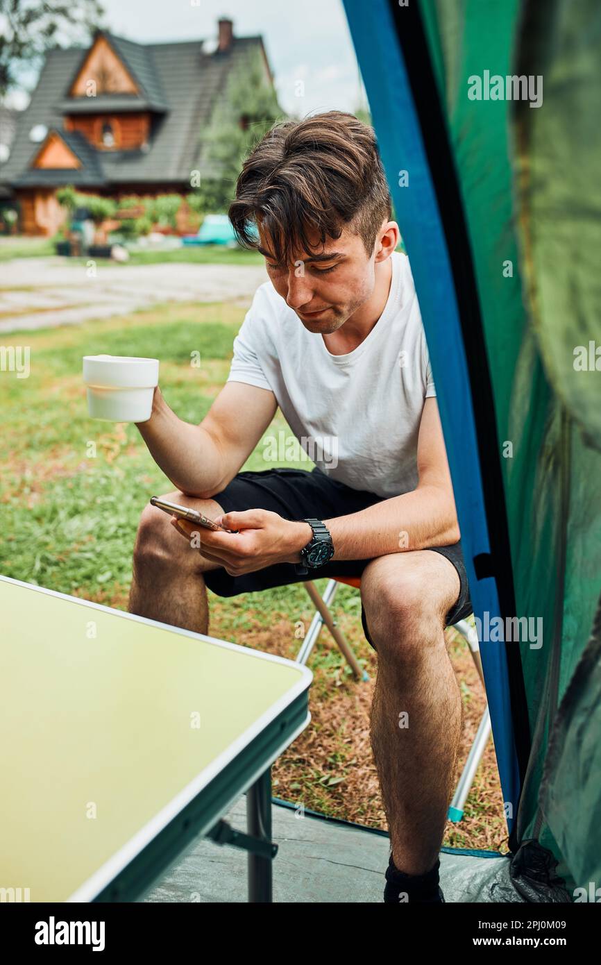 Un jeune homme buvant un café assis devant la tente le matin. Adolescent profitant de temps libre pendant le week-end de voyage en été Banque D'Images