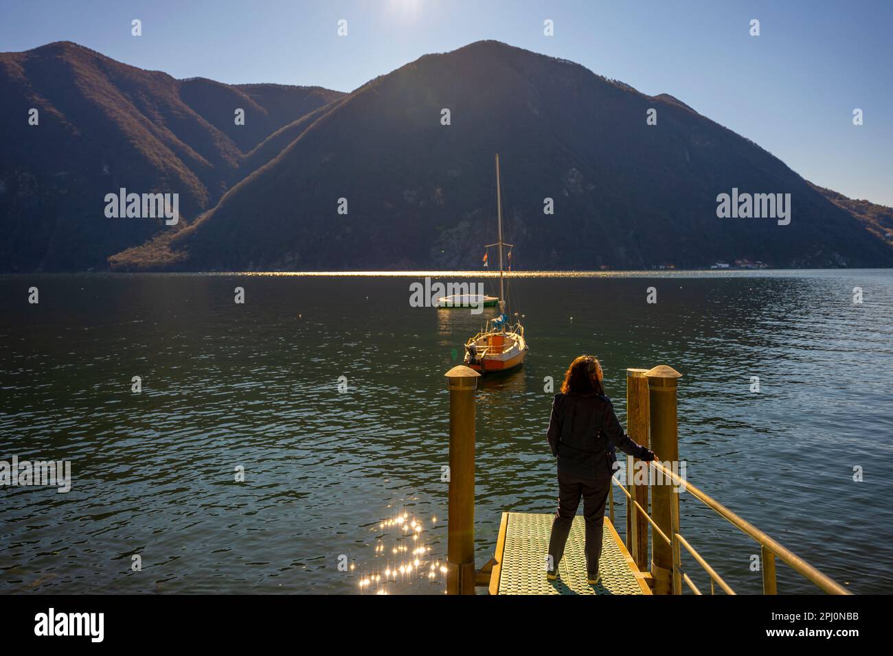 Femme debout sur un quai avec un Voilier sur le lac de Lugano et chaîne de montagnes en une journée ensoleillée avec ciel clair à Lugano, Tessin, Suisse. Banque D'Images