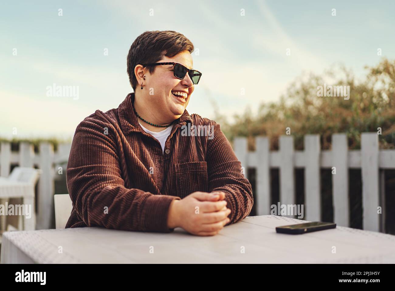 Personne non binaire souriante de plus grande taille avec des lunettes de  soleil dans un café extérieur, en regardant loin. Veste en velours marron,  fond bleu ciel Photo Stock - Alamy