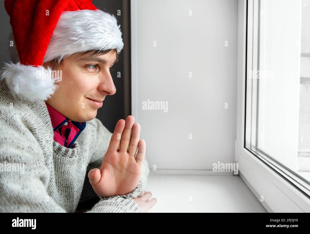 Joyeux jeune homme dans le chapeau du Père Noël vague avec une main près de la fenêtre dans la salle Banque D'Images