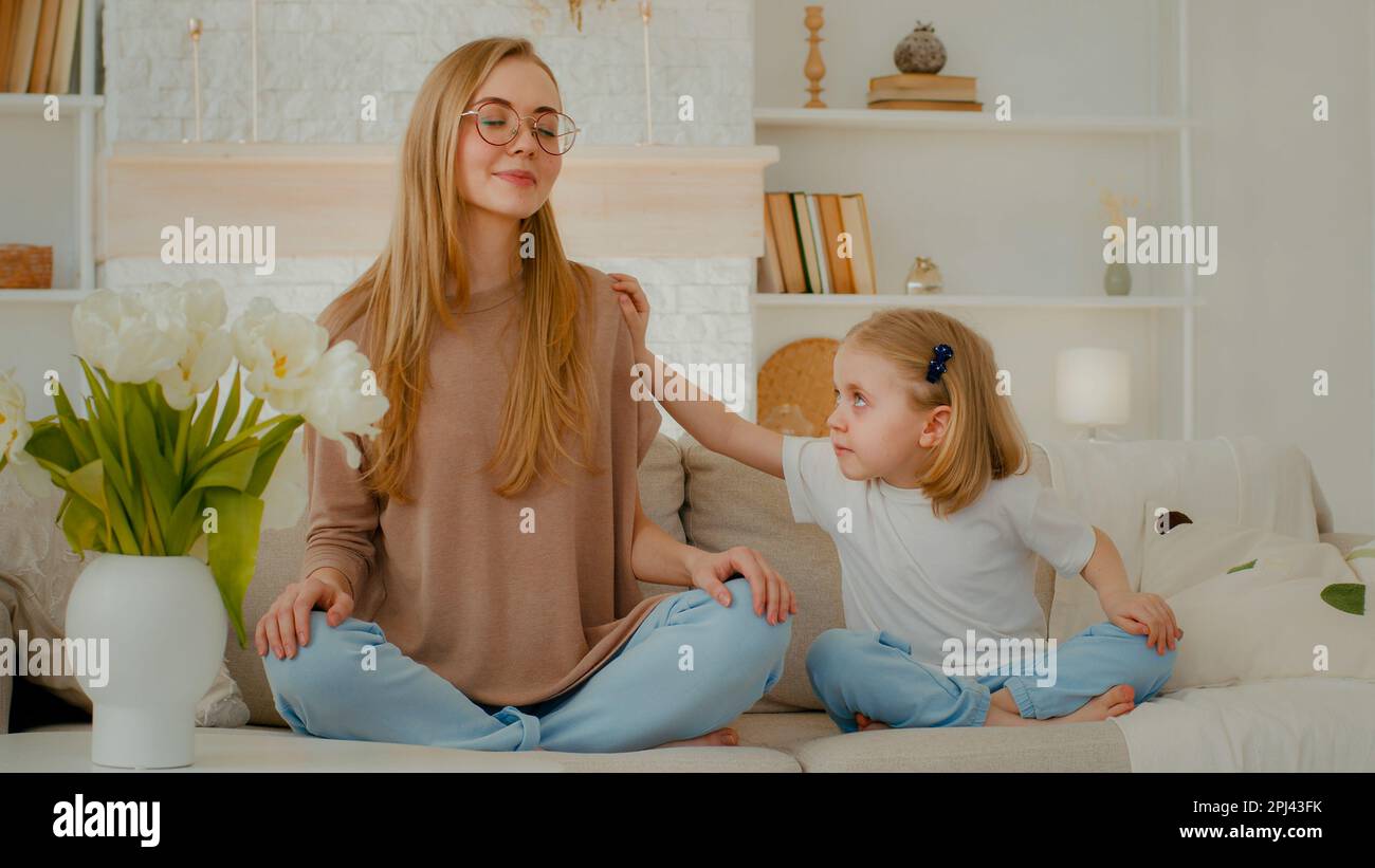 Conscient de la mère caucasienne calme femme méditant avec les yeux fermés dans la position de lotus faisant yoga exercice méditation asseyez-vous sur le canapé à la maison petite fille d'enfant Banque D'Images
