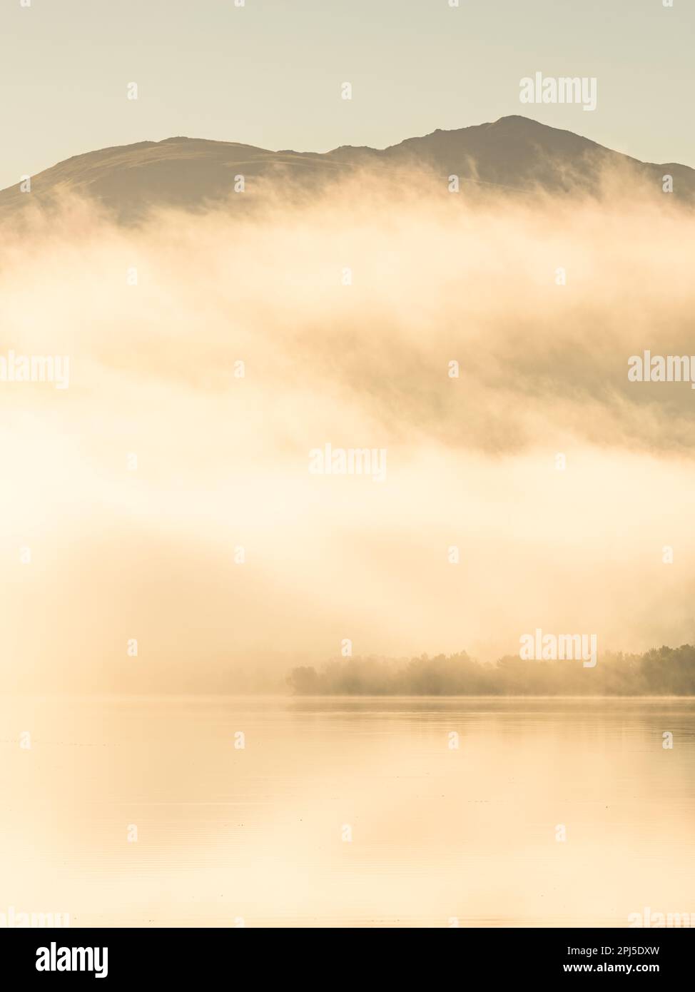 Une paisible scène matinale d'un lac brumeux reflétant les montagnes ensoleillées et les arbres, entouré par la nature intacte. Banque D'Images