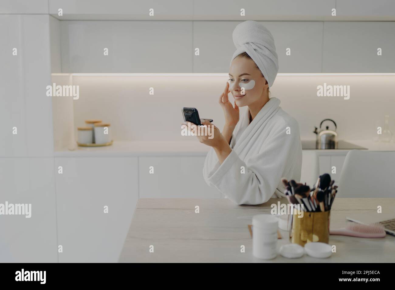 Jeune femme attrayante en peignoir et serviette turban sur la tête appliquant des taches cosmétiques sous les yeux des cercles sombres, regardant dans un miroir compact et smil Banque D'Images
