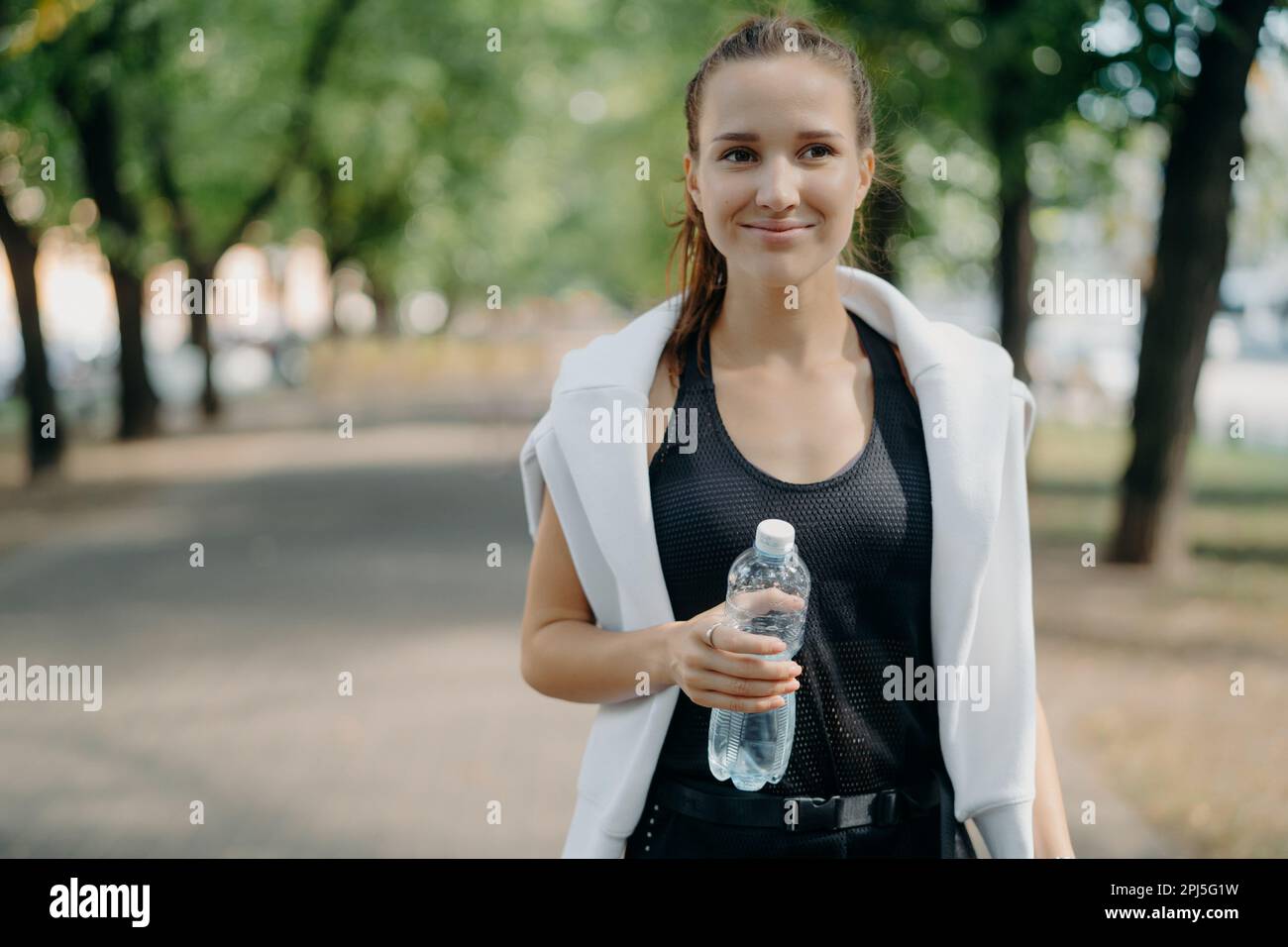 Une jeune femme gaie tient une bouteille d'eau en plein air en tenue active prend une pause après que les promenades d'entraînement dans le parc se rafraichissement après le jogging fe Banque D'Images
