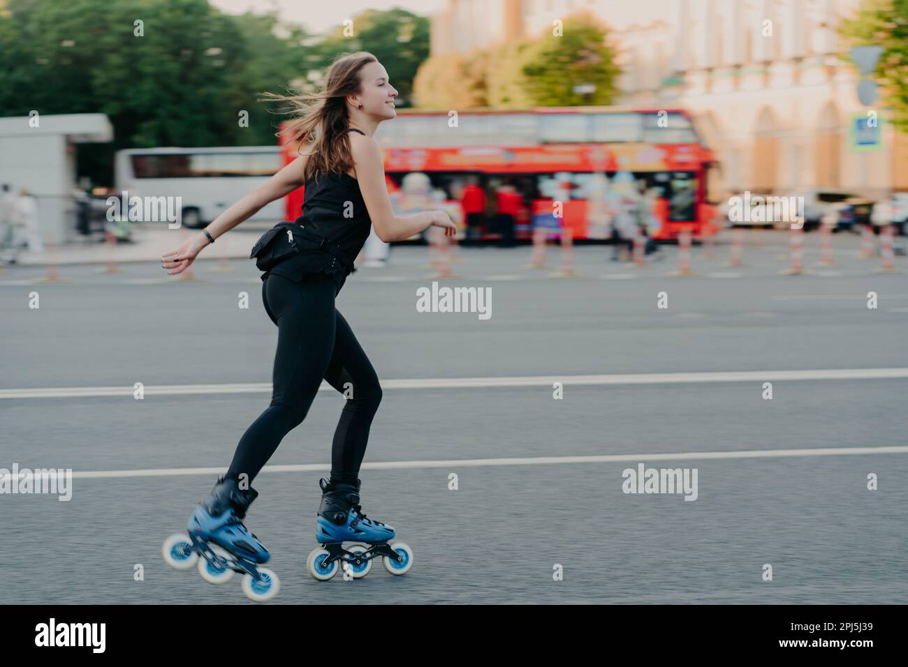 Photo en extérieur d'une jeune femme active mince aime le roller pendant le temps libre vêtu de vêtements de noirceur pose en milieu urbain sur la route contre le flou Banque D'Images