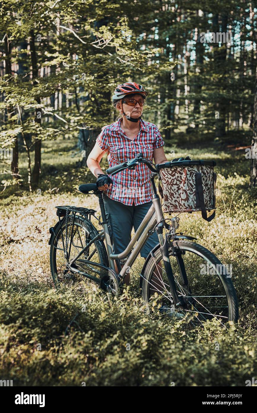 Femme active qui passe des vacances d'été gratuites lors d'un voyage à vélo dans une forêt. Femme portant un casque de vélo et des gants tenant un vélo avec un panier Banque D'Images