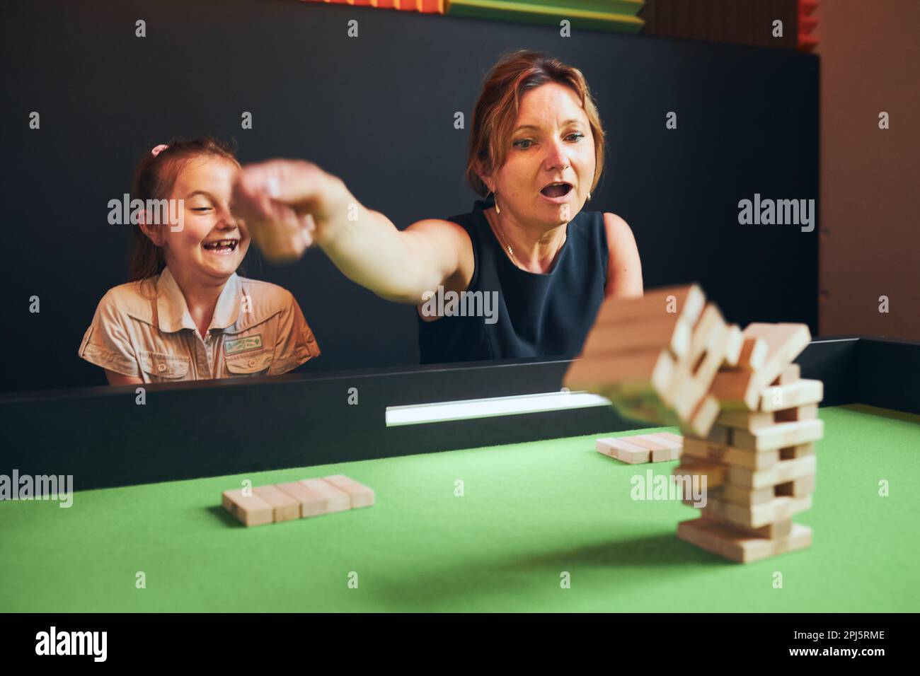 Une femme excitée jouant à jenga avec sa fille dans la salle de jeux. Femme qui retire le bloc de la pile. Tour de chute de blocs de bois. Jeu de compétence et de fu Banque D'Images