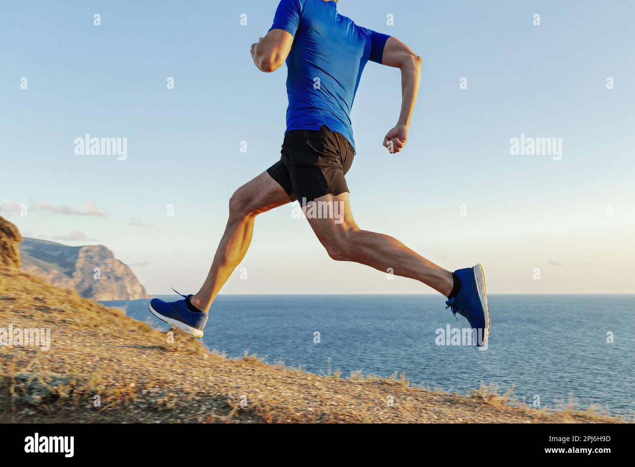 le coureur de sexe masculin monte en côte, fait un sentier maritime sur gazon fané, fait une course de cross-country, un maillot bleu et un short noir Banque D'Images