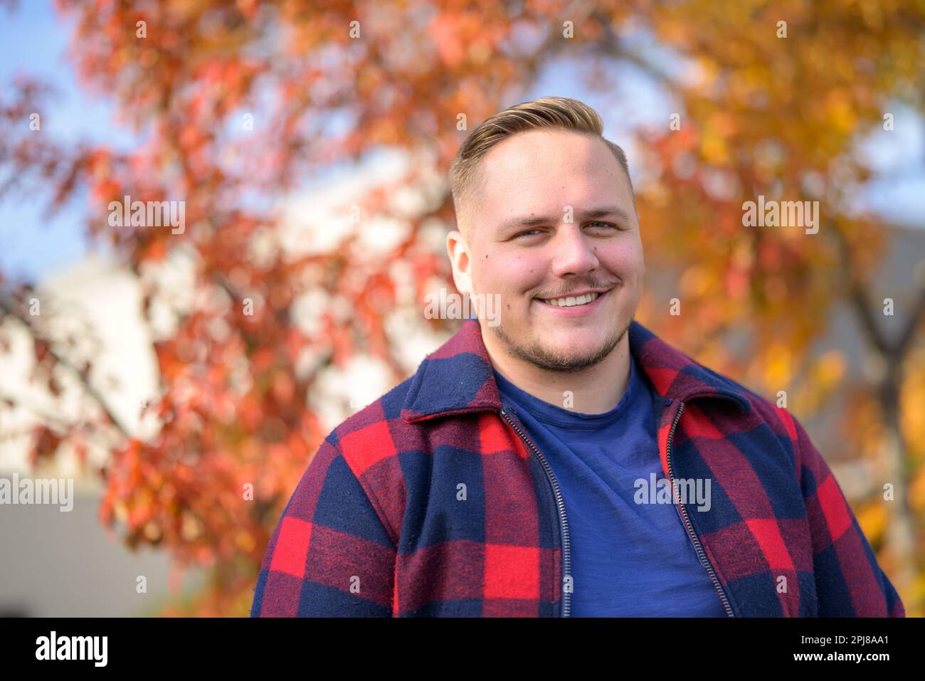 Vue de face d'un jeune homme avec un sourire éclatant dans une veste paysanne se dresse devant un fond d'automne avec des arbres et des feuilles jaunes Banque D'Images