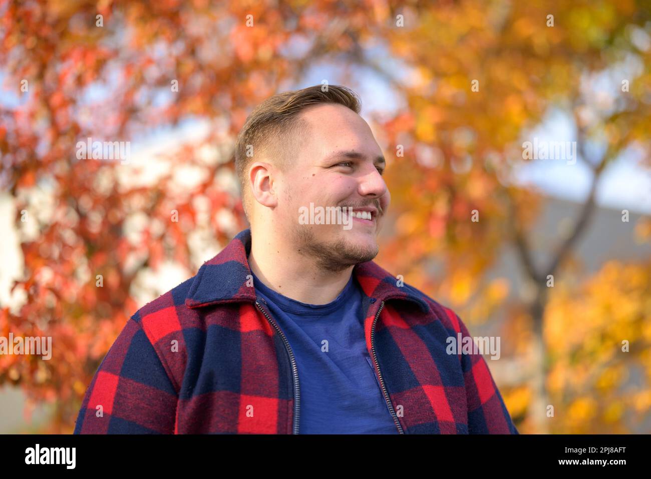 Un jeune homme avec un sourire éclatant dans une veste paysanne se tient devant un fond d'automne avec des arbres et des feuilles jaunes Banque D'Images