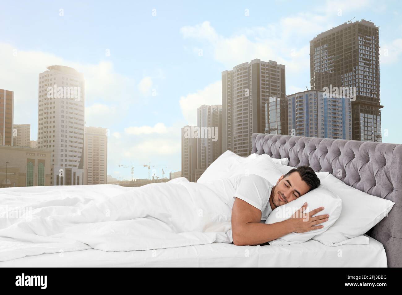 Homme dormant sur un lit avec des oreillers moelleux et une vue magnifique sur le paysage urbain en arrière-plan. Bonne nuit de sommeil malgré l'agitation urbaine Banque D'Images