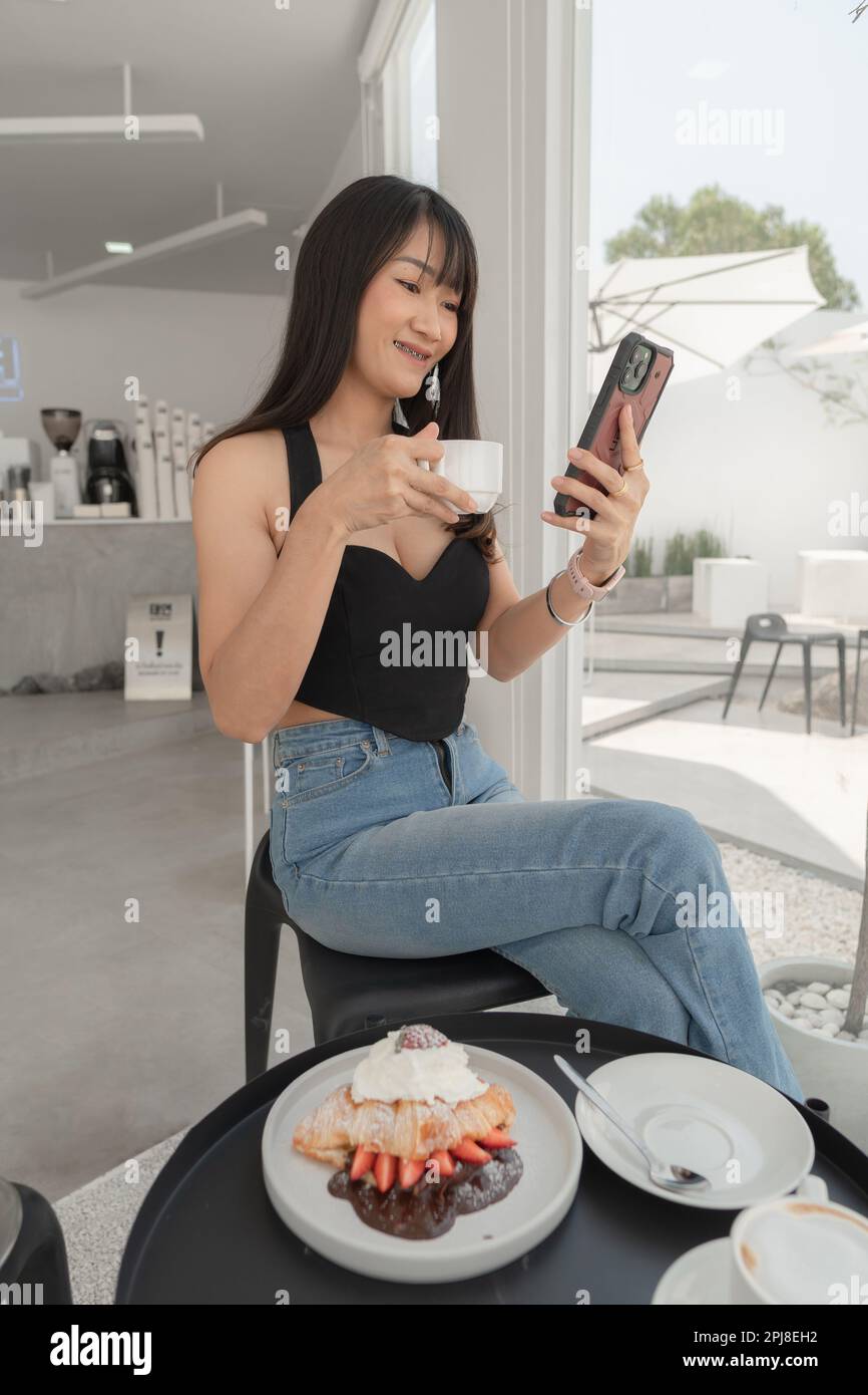 Belle femme asiatique sexy en robe noire boire du café et jouer smartphone dans le café, blanc minimaliste style. Banque D'Images