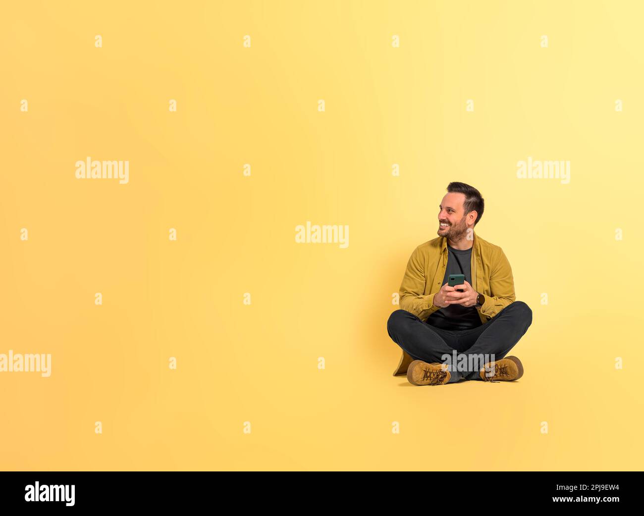 Pleine longueur de gaie bonne apparence homme moyen de messagerie texte sur le smartphone et regardant loin en étant assis avec les jambes croisées isolées sur jaune Banque D'Images