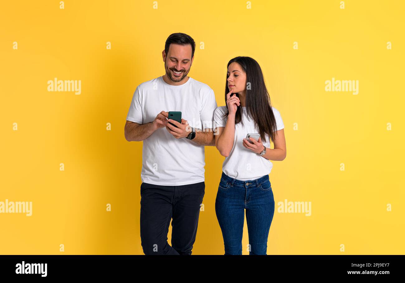 Curieuse jeune femme adulte avec la main sur le menton pélécher dans le téléphone mobile de l'ami et essayant de lire ses messages sur fond jaune Banque D'Images