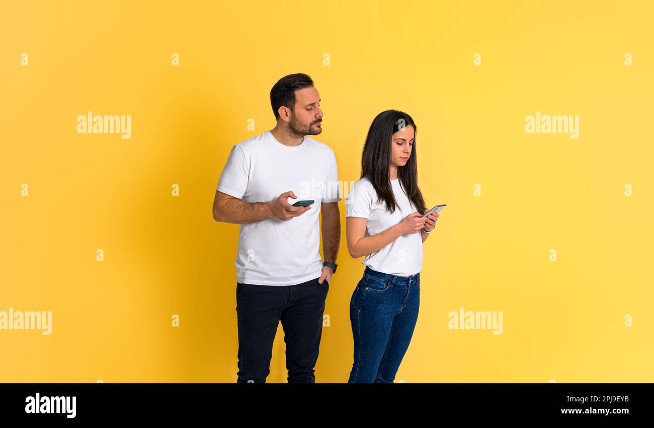 Jeune homme adulte possessif qui se parait sur le smartphone de sa petite amie et qui essaie de lire ses messages texte tout en étant isolé sur fond jaune Banque D'Images