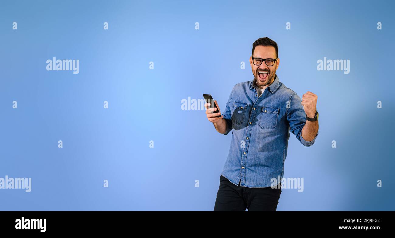 Un homme de taille moyenne enthousiaste portant une chemise en denim et des lunettes avec un téléphone intelligent qui tire le poing et crie joyeusement sur fond bleu Banque D'Images