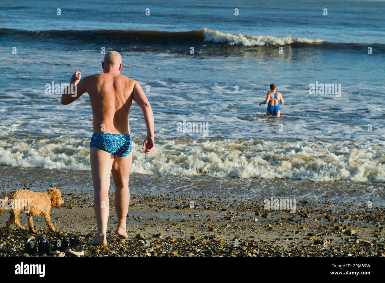 Homme dans les trunks courant dans la mer avec chien sur Avon Beach Christchurch, Royaume-Uni en hiver Banque D'Images