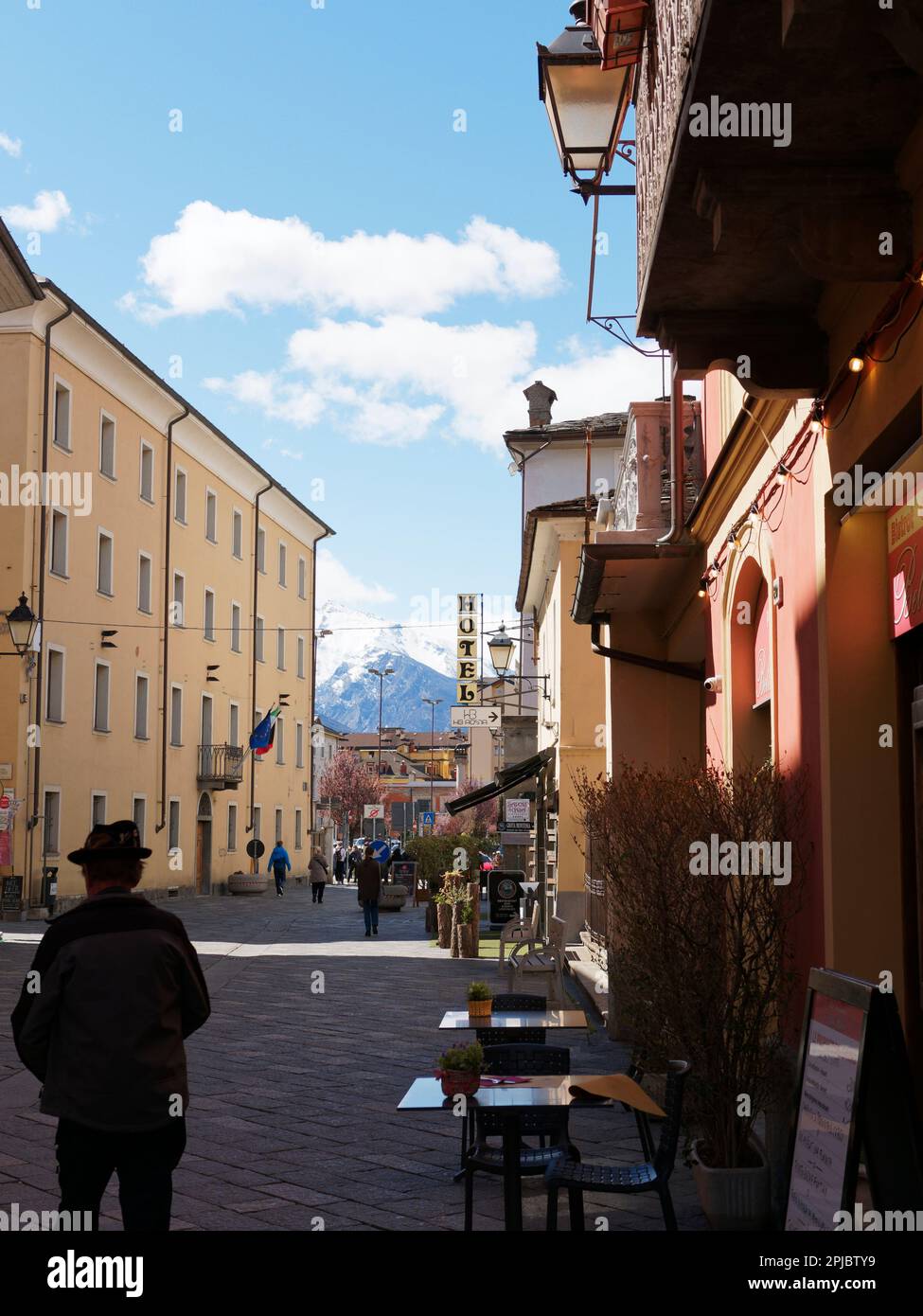 Rue d'Aoste avec un homme portant un chapeau en silhouette et des tables et chaises de café/bar en premier plan et une montagne au loin, vallée d'Aoste, Italie Banque D'Images