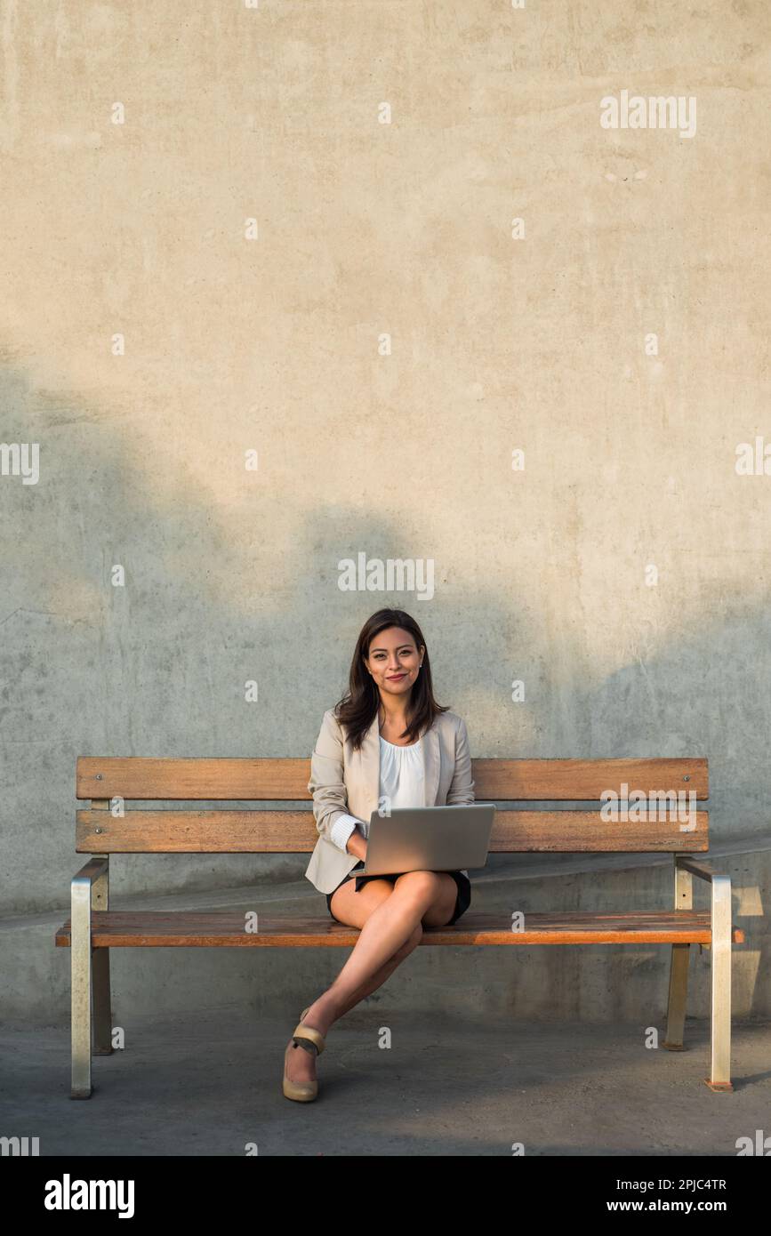 Image verticale d'une femme d'affaires Latina assise sur un banc regardant l'appareil photo et tenant un ordinateur personnel. Banque D'Images