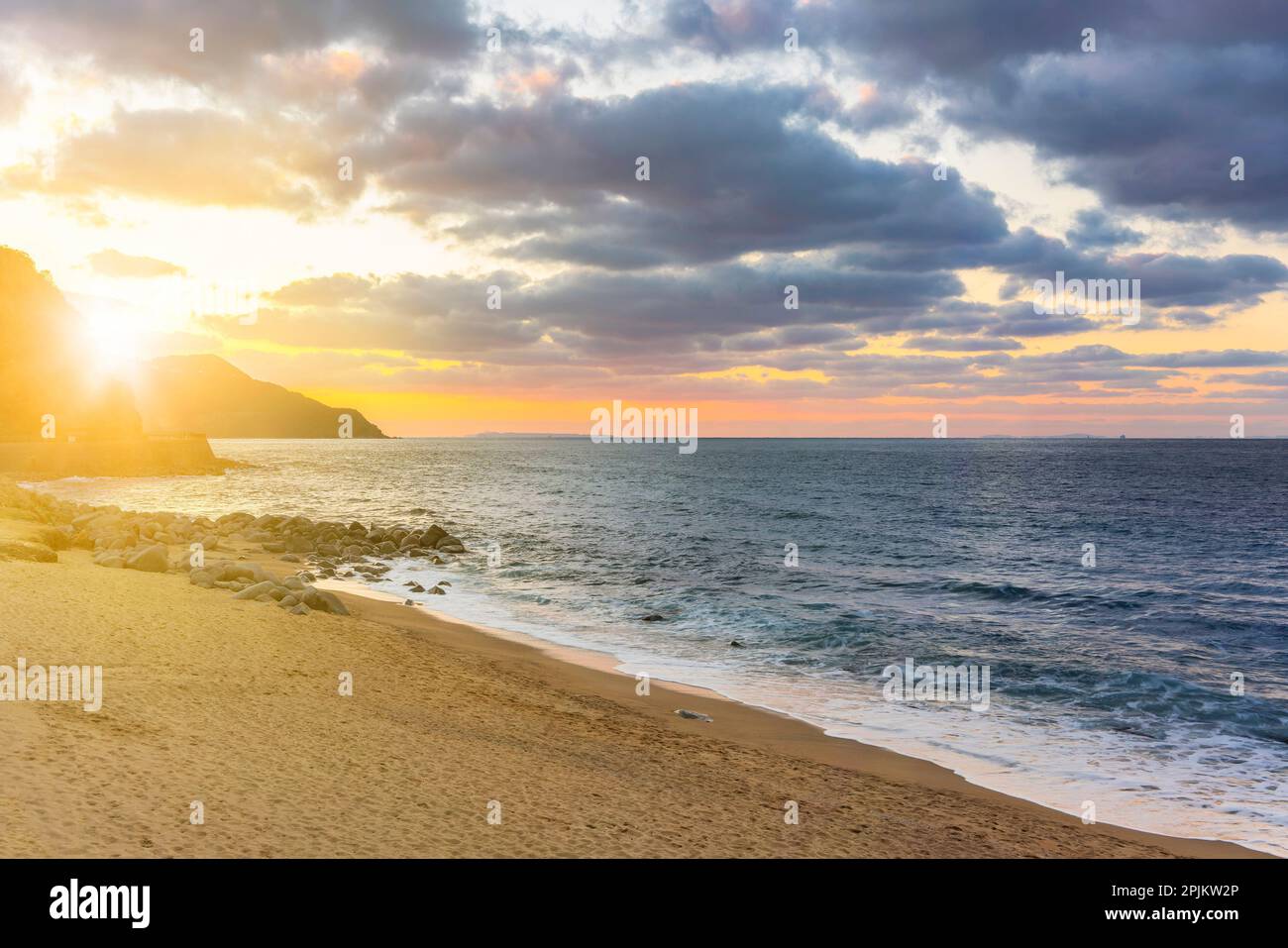 Lumière du coucher de soleil aux couleurs or et rose qui filtre à travers les nuages au-dessus de la silhouette d'une côte avec en premier plan une plage de sable avec des rochers et Banque D'Images