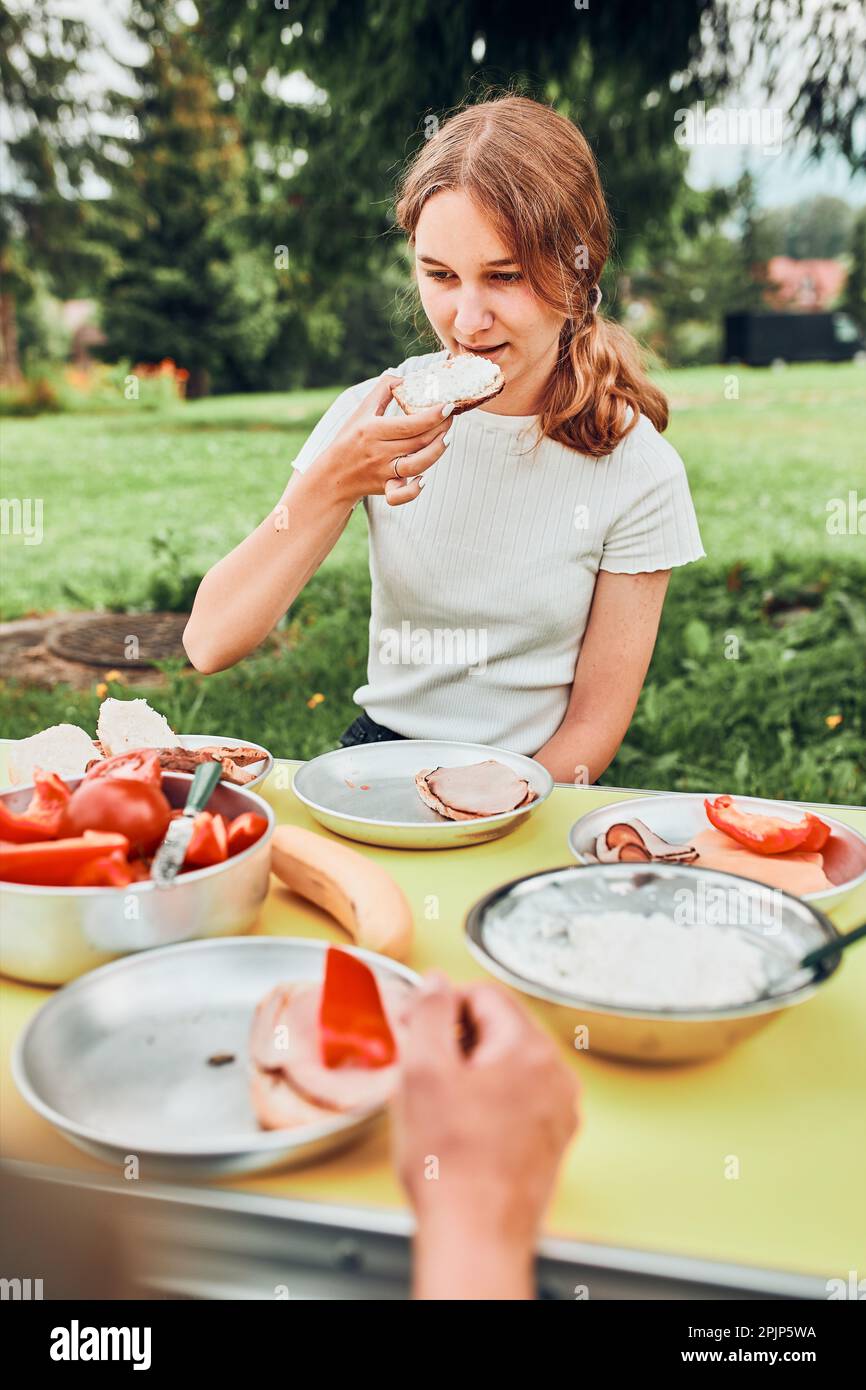 Famille prenant le petit déjeuner à l'extérieur sur le camping pendant les vacances d'été. Pain, fromage cottage, viande froide, tomates, fruits et tasses à café sur table Banque D'Images