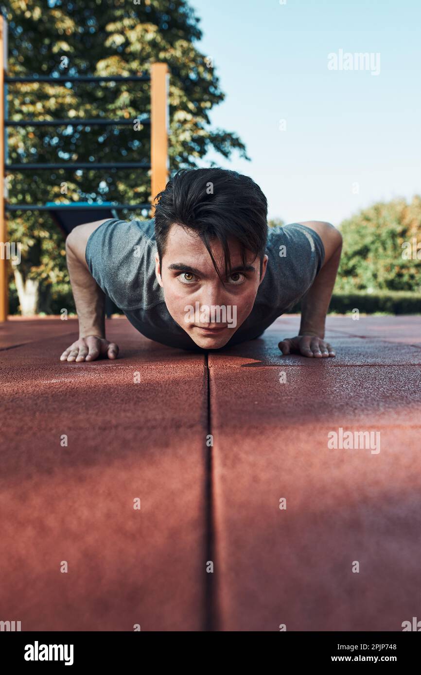 Jeune homme faisant des push-up sur un sol en caoutchouc rouge pendant son entraînement dans un parc d'entraînement moderne de rue de calisthéniques. Homme portant des vêtements de sport regardant la caméra Banque D'Images