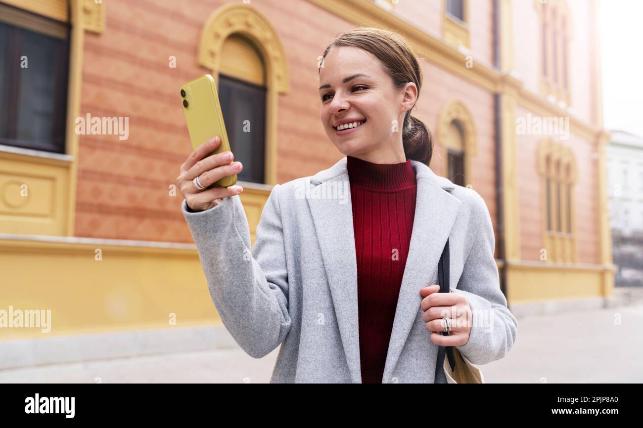 Jeune femme élégante portant un manteau lisant un message sur son téléphone mobile tout en marchant dans la rue et en souriant. Banque D'Images