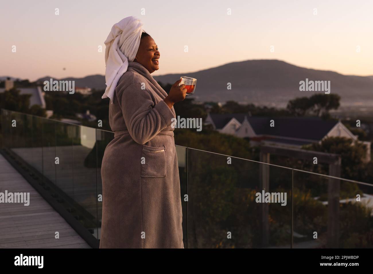 Femme afro-américaine de bonne taille, portant un peignoir et une serviette sur la tête, buvant du thé sur le balcon Banque D'Images