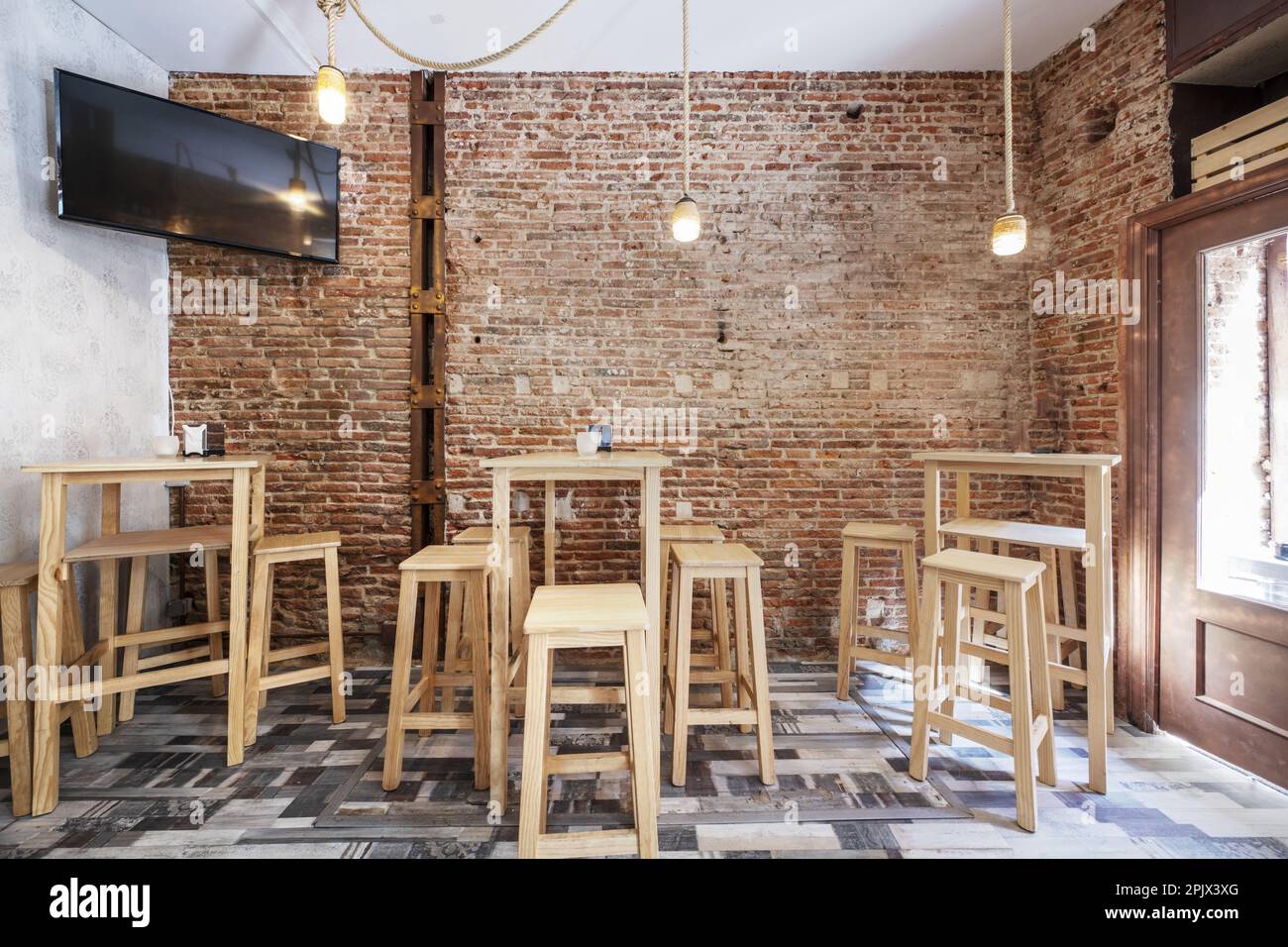 Un bar vide avec des murs en briques brutes et de grandes tables en bois blanc avec des tabourets assortis, un sol en mosaïque de bois et une télévision Banque D'Images