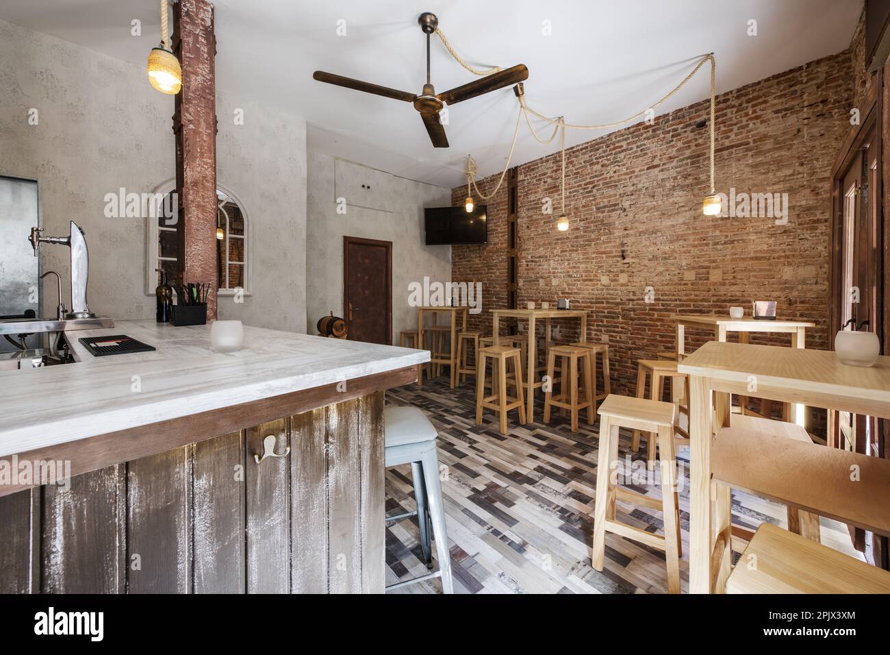 Un bar vide avec un comptoir en bois blanc, un ventilateur de plafond, des murs en briques brutes et de grandes tables en bois blanc avec des tabourets assortis, un plancher en mosaïque de bois et Banque D'Images