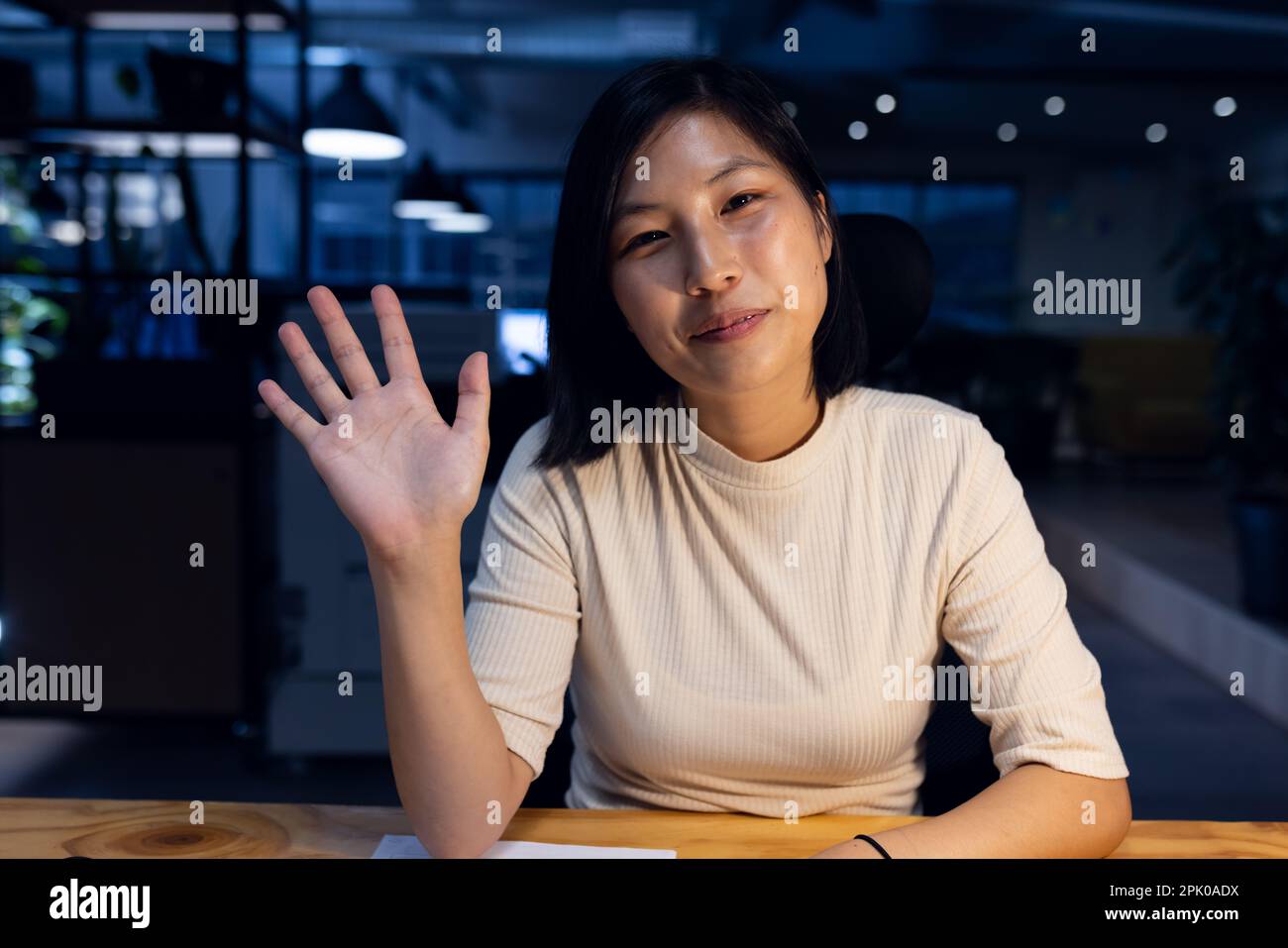 Bonne femme d'affaires asiatique assise et agitant, faisant un appel vidéo, travaillant tard au bureau Banque D'Images