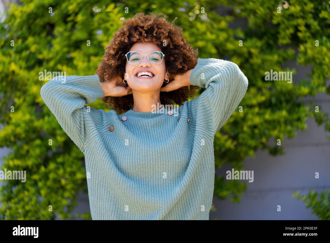 Portrait de jeune femme gaie biraciale avec les mains dans les cheveux afro souriant et debout dans la cour Banque D'Images