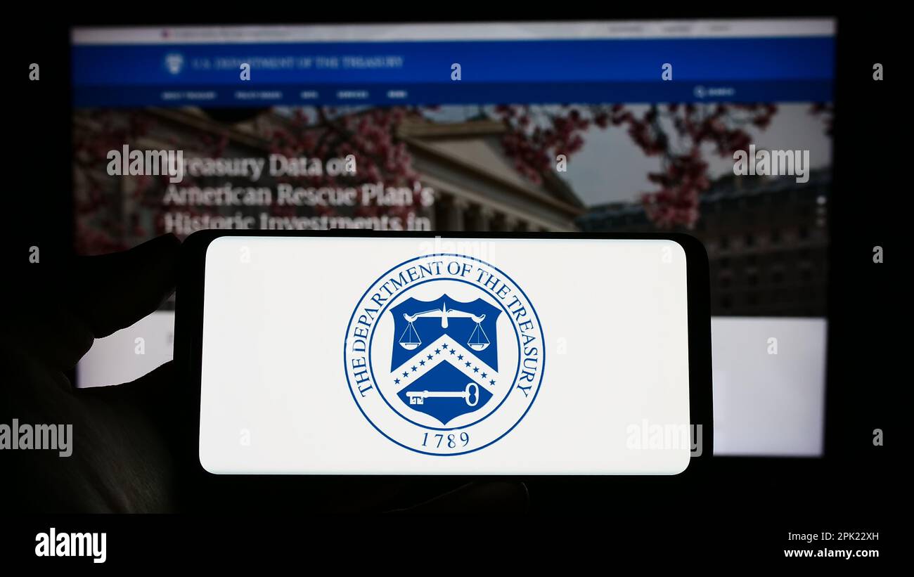 Personne tenant un téléphone portable avec le sceau du Département du Trésor des États-Unis (USDT) à l'écran devant la page Web. Mise au point sur l'affichage du téléphone. Banque D'Images