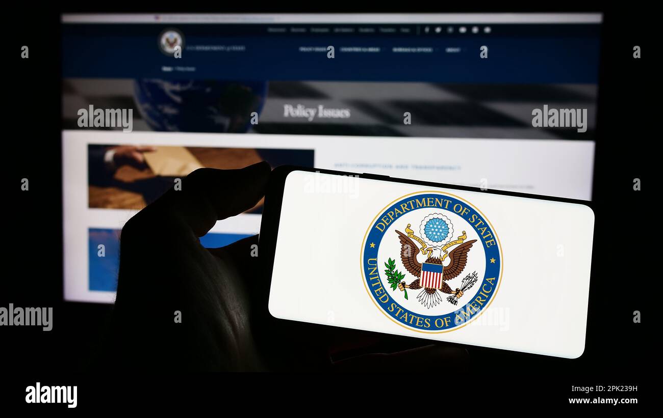 Personne tenant un téléphone portable avec le sceau du Département d'État des États-Unis (DOS) sur l'écran en face de la page Web. Mise au point sur l'affichage du téléphone. Banque D'Images