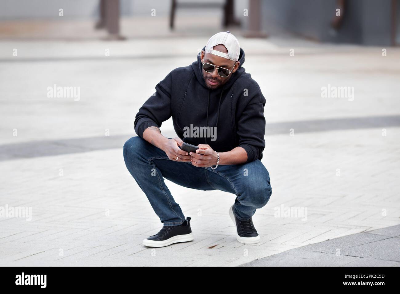 Cool African American homme utilisant un smartphone, s'accroupir dans la rue de la ville, porte noir sweat à capuche, jeans et baskets, casquette de baseball et lunettes de soleil. Style Banque D'Images