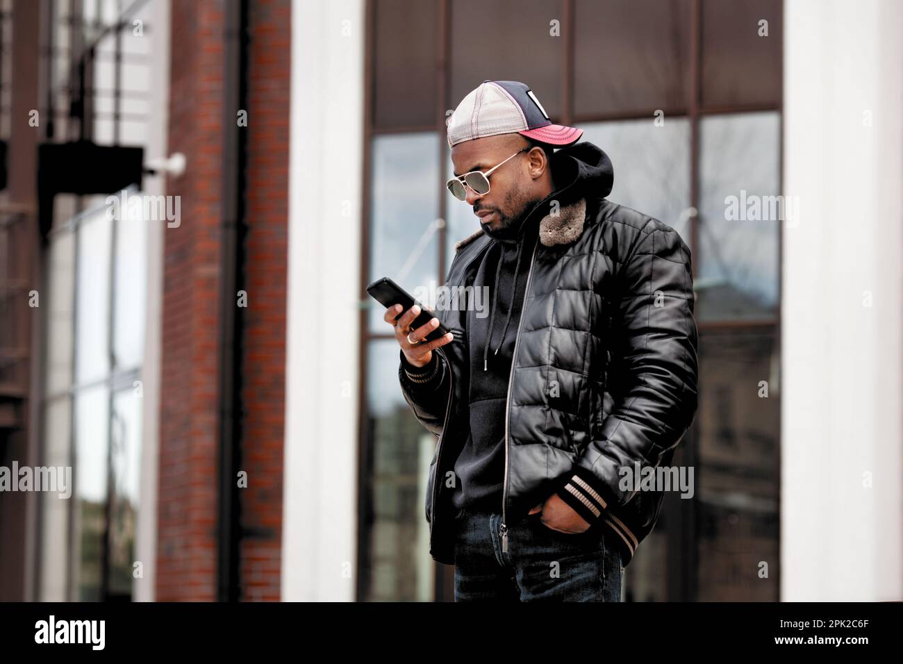 Cool African American Guy utilise un smartphone, se tient dans la rue de la ville après le bâtiment, porte une veste en cuir, casquette de baseball et des lunettes de soleil. Look homme élégant Banque D'Images