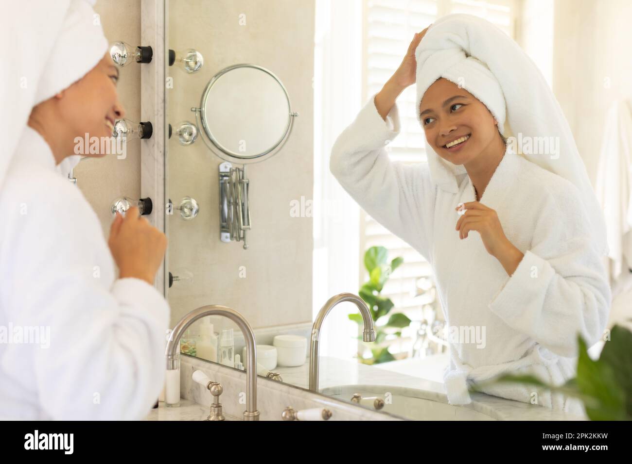 Bonne femme asiatique portant une serviette sur la tête et regardant dans le miroir de salle de bains Banque D'Images