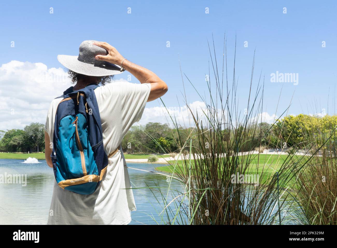 Vue arrière d'un homme avec sac à dos, chapeau de soleil et t-shirt observe un lagon au milieu de la forêt lors d'une randonnée dans la nature tropicale au Mexique Banque D'Images