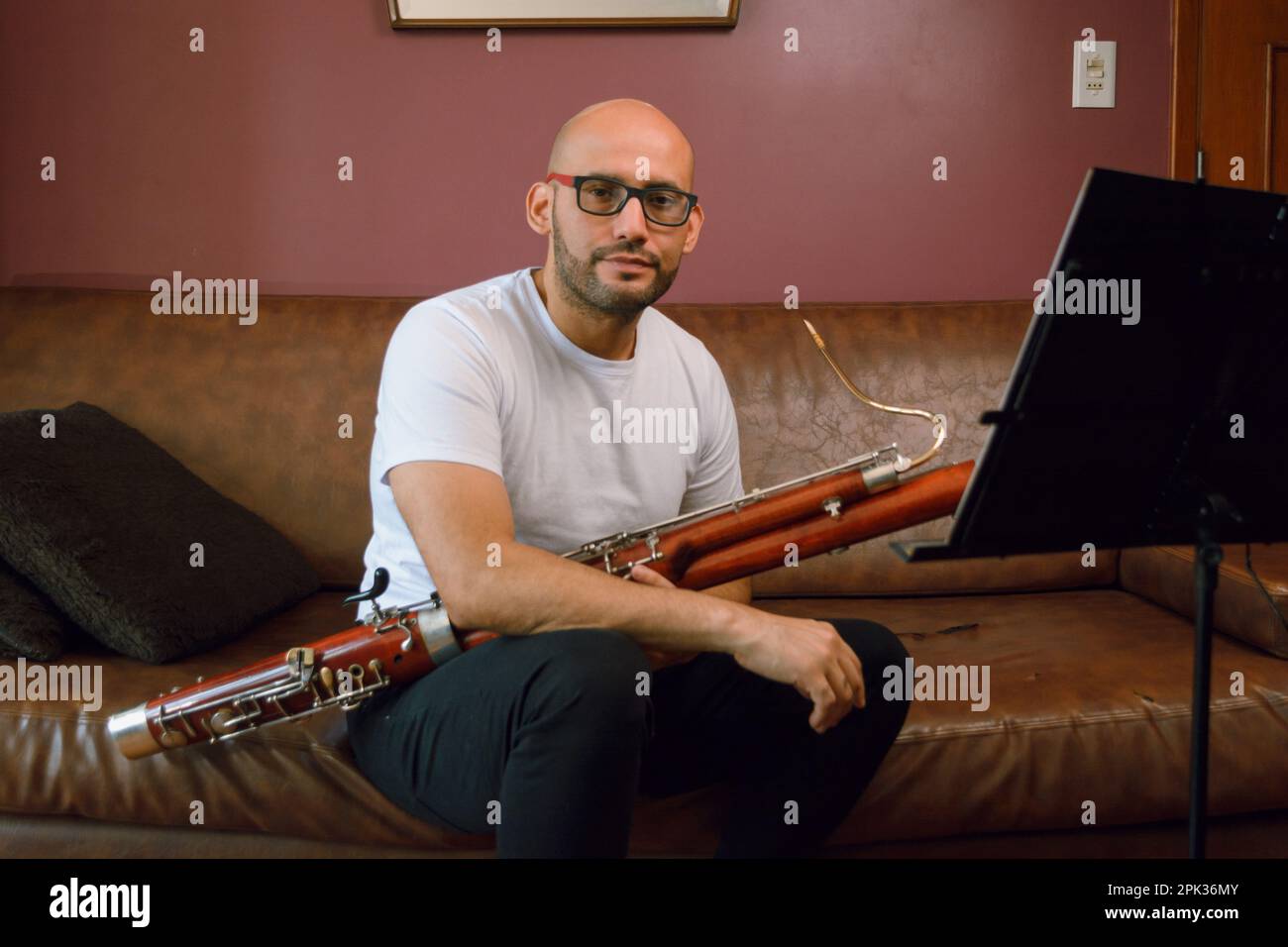 portrait d'un jeune homme de latino bald avec des lunettes tenant un basson et un stand de musique assis sur un canapé en cuir marron dans le salon qui regarde le c Banque D'Images