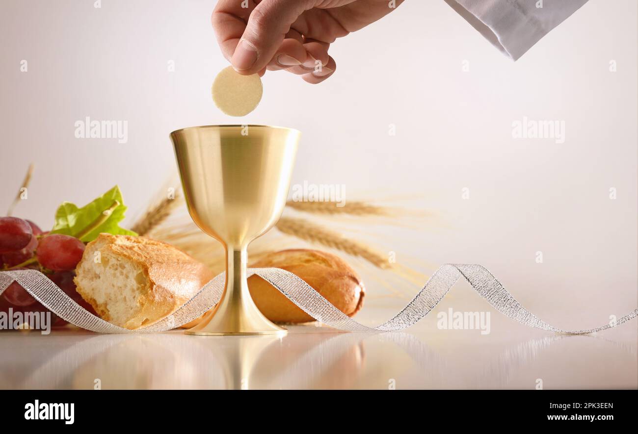 Eucharistie avec la main du prêtre tenant une hôte et une tasse de vin consacré sur une table avec du pain et des raisins et un fond isolé. Vue avant. Banque D'Images