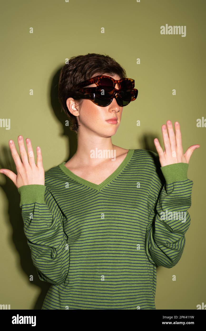 brunette femme en pull rayé portant plusieurs lunettes de soleil tendance sur fond vert, image de stock Banque D'Images