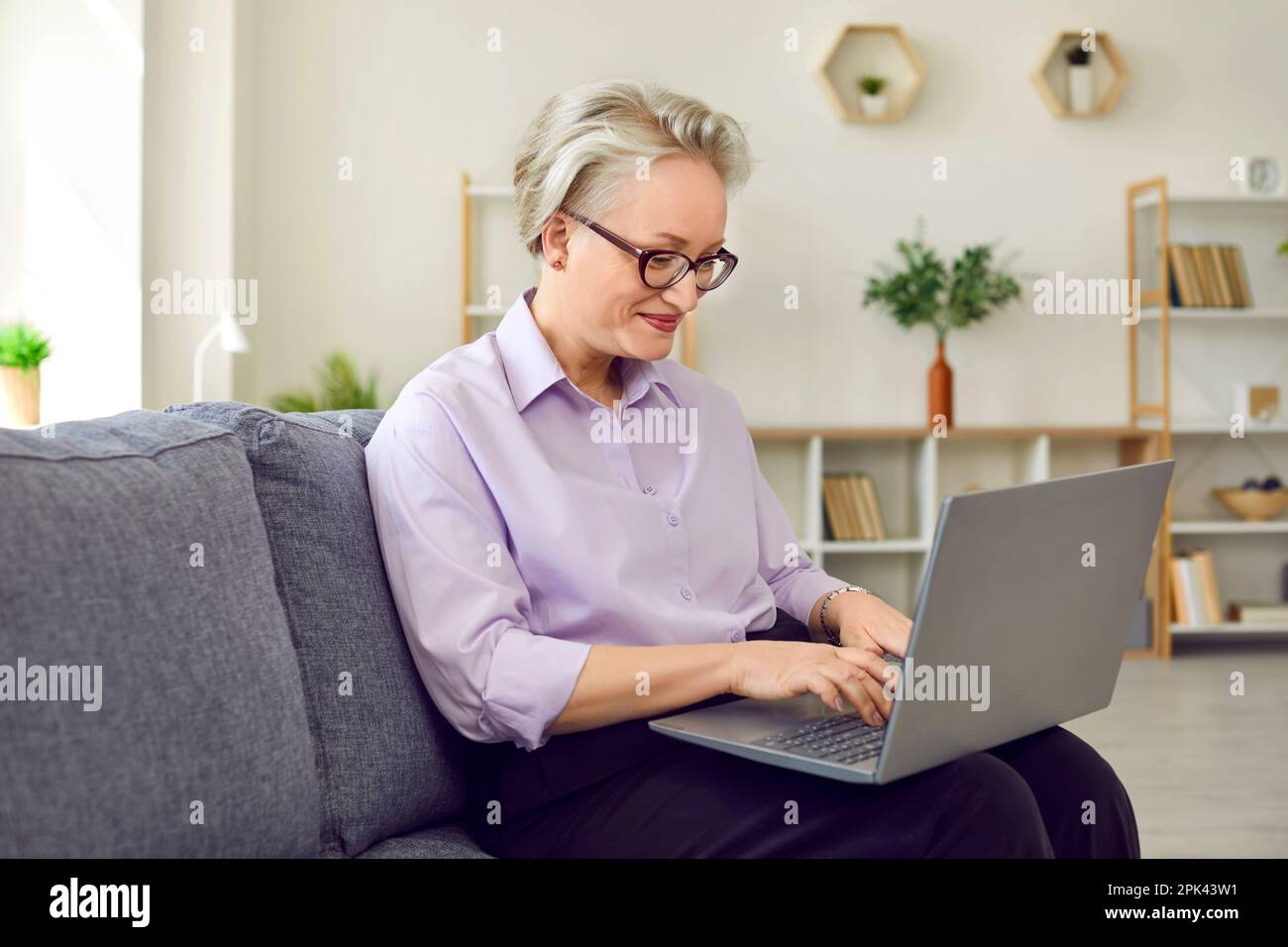 Une femme mûre en lunettes travaille à taper un ordinateur portable assis sur un canapé dans son bureau confortable. Banque D'Images