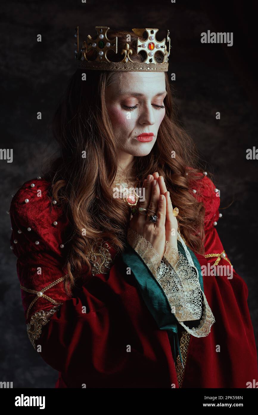 reine médiévale en robe rouge avec couronne priant sur fond gris foncé. Banque D'Images