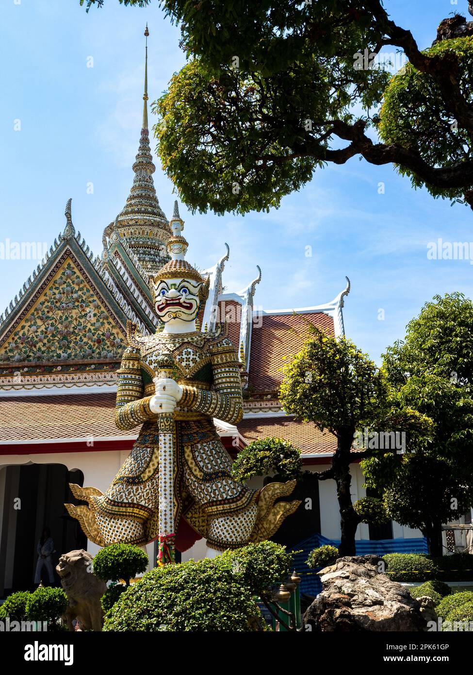 Statue de gardien de démon géant colorée au temple Wat Arun de Bangkok, en Thaïlande, entourée d'arbres verts luxuriants et d'un ciel bleu éclatant. La statue respire un Banque D'Images