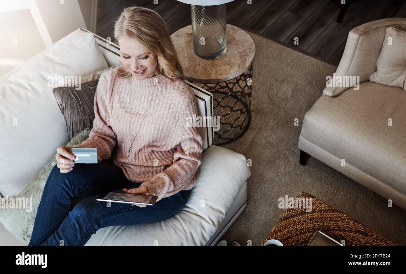 Shopping et confort, le pari des deux mondes. une femme mûre utilisant une carte de crédit et une tablette numérique tout en se relaxant à la maison. Banque D'Images