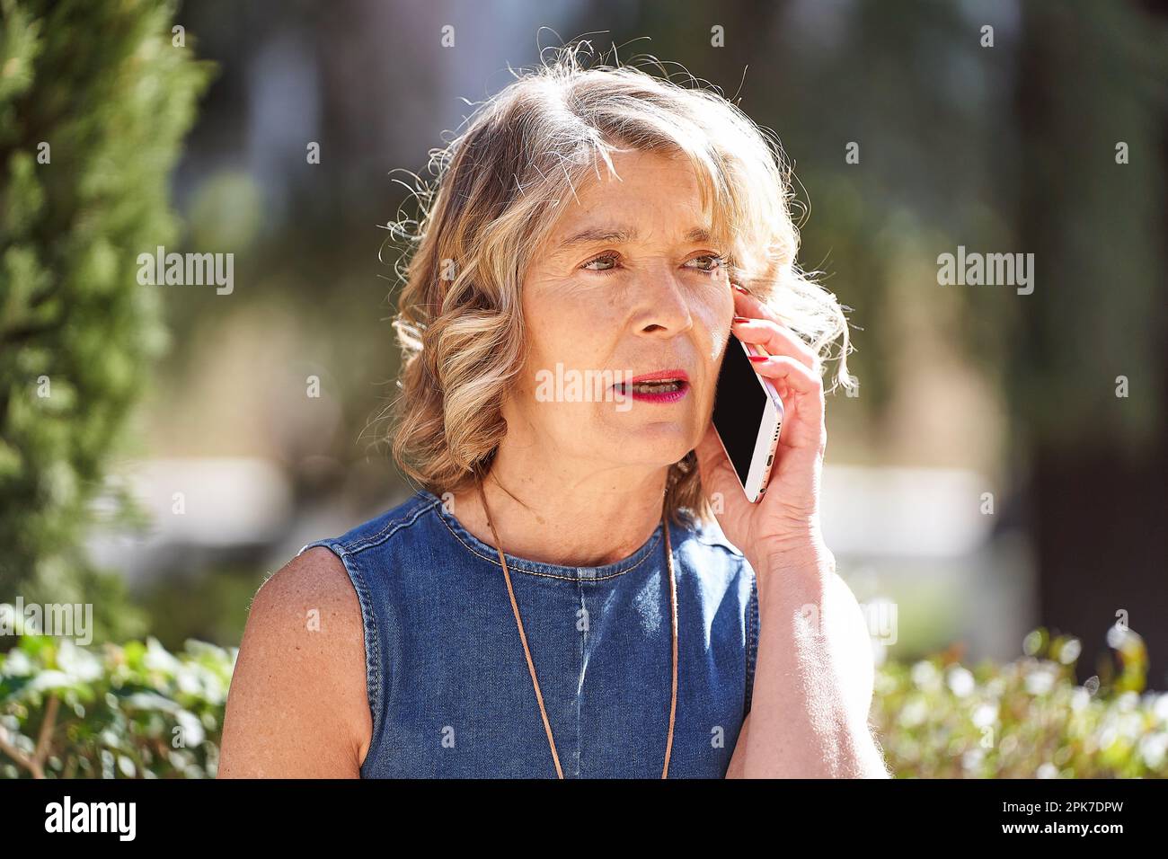 Une femme dans ses 60s chats au téléphone pendant une pause en plein air : une femme prend une pause de ses activités quotidiennes et discute avec quelqu'un sur son téléphone portable tout en étant assise sur un banc extérieur. Banque D'Images