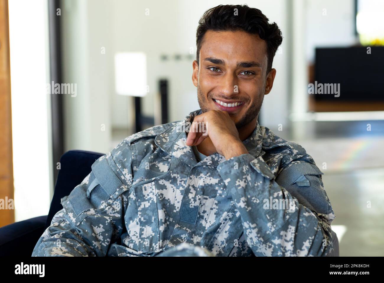 Portrait d'un homme biracial heureux portant un uniforme militaire, assis dans un fauteuil à la maison. Style de vie, service militaire et temps libre, sans modification. Banque D'Images