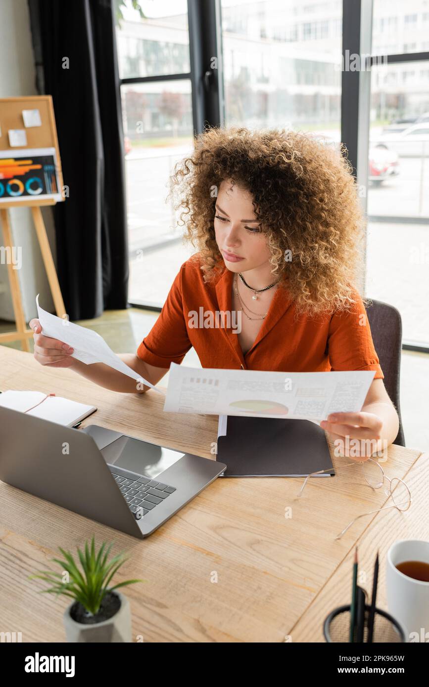 femme d'affaires maudite regardant des infographies près d'un ordinateur portable et une tasse de thé sur le bureau, image de stock Banque D'Images