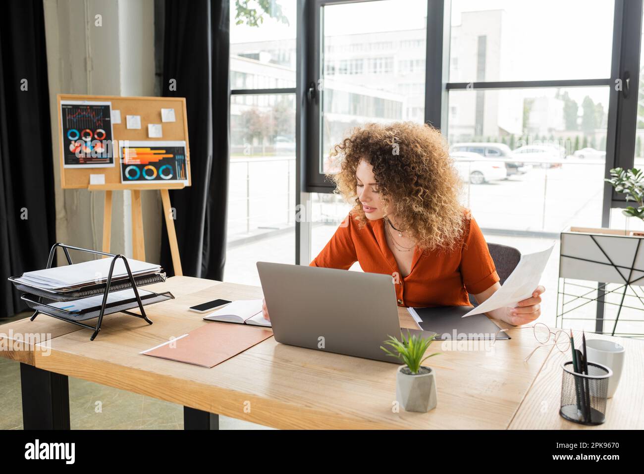 femme d'affaires maudite tenant des infographies et utilisant un ordinateur portable et une tasse de thé sur le bureau, image de stock Banque D'Images