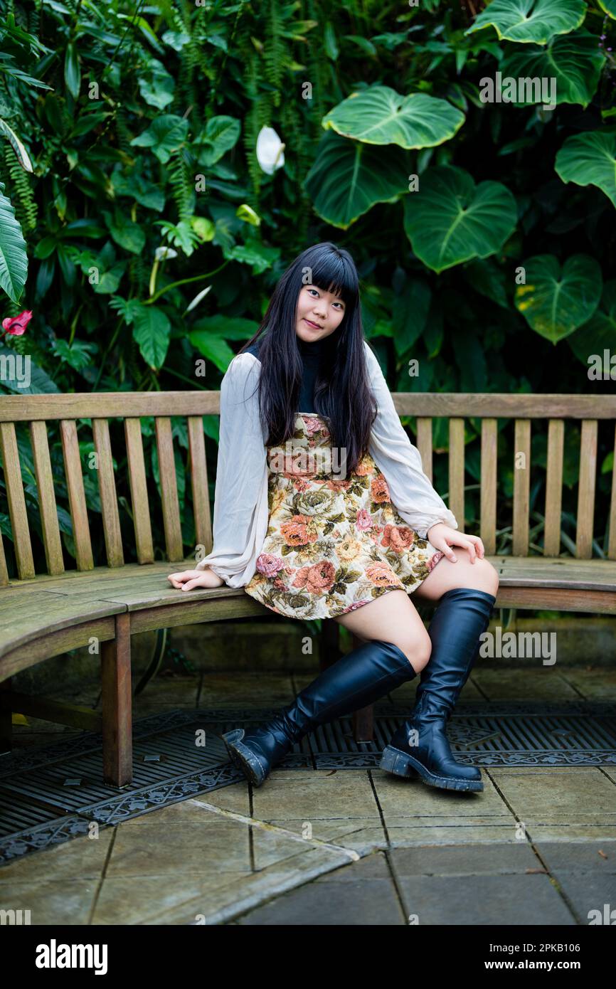 Jeune femme asiatique assise sur un banc de teck dans un jardin victorien | jambes en Z | sourire | tons chauds | bottes hauteur genou | robe courte Banque D'Images